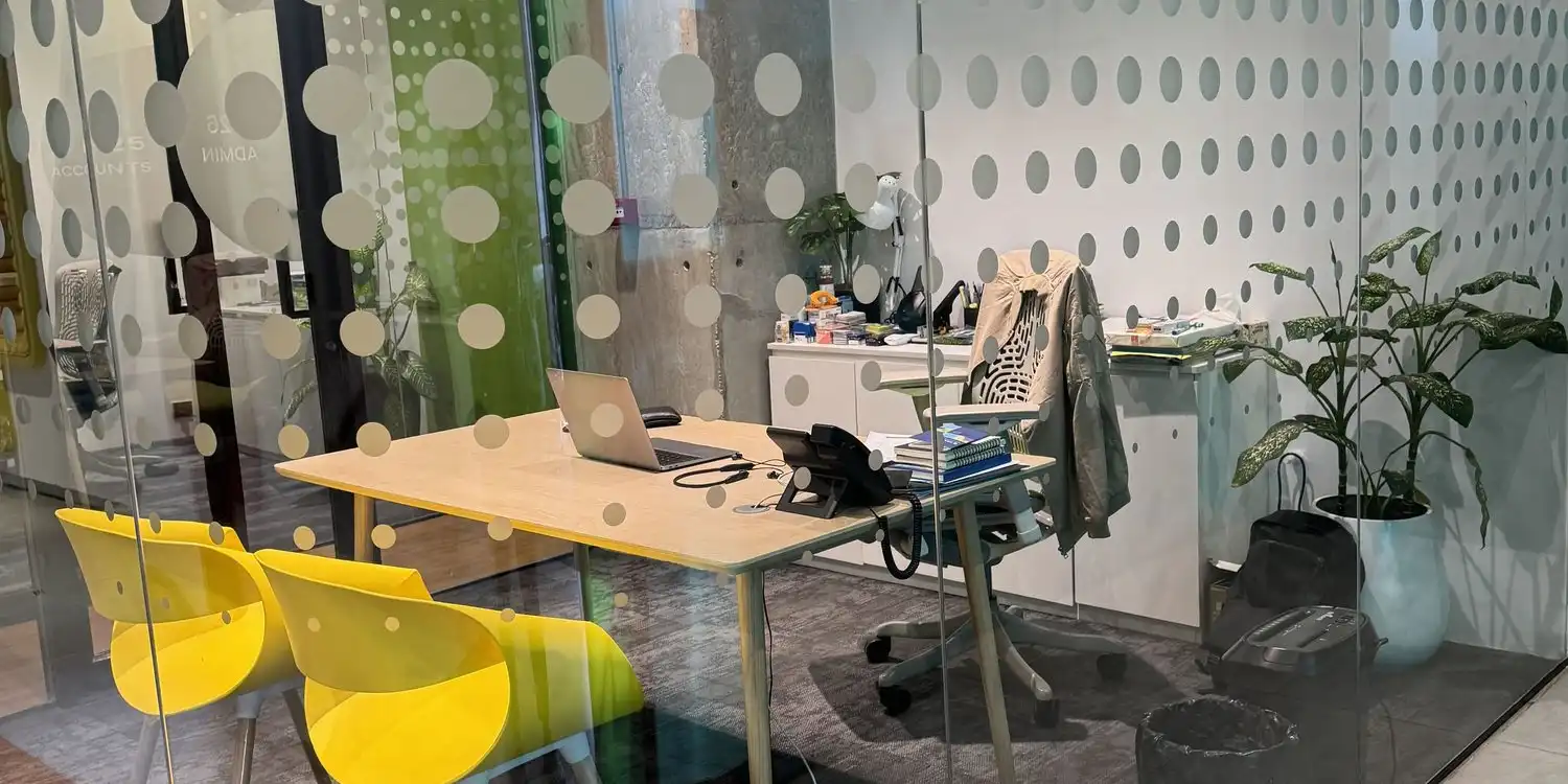 A glass-walled office with a desk, an open laptop, two yellow chairs and potted plants