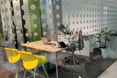 A glass-walled office with a desk, an open laptop, two yellow chairs and potted plants