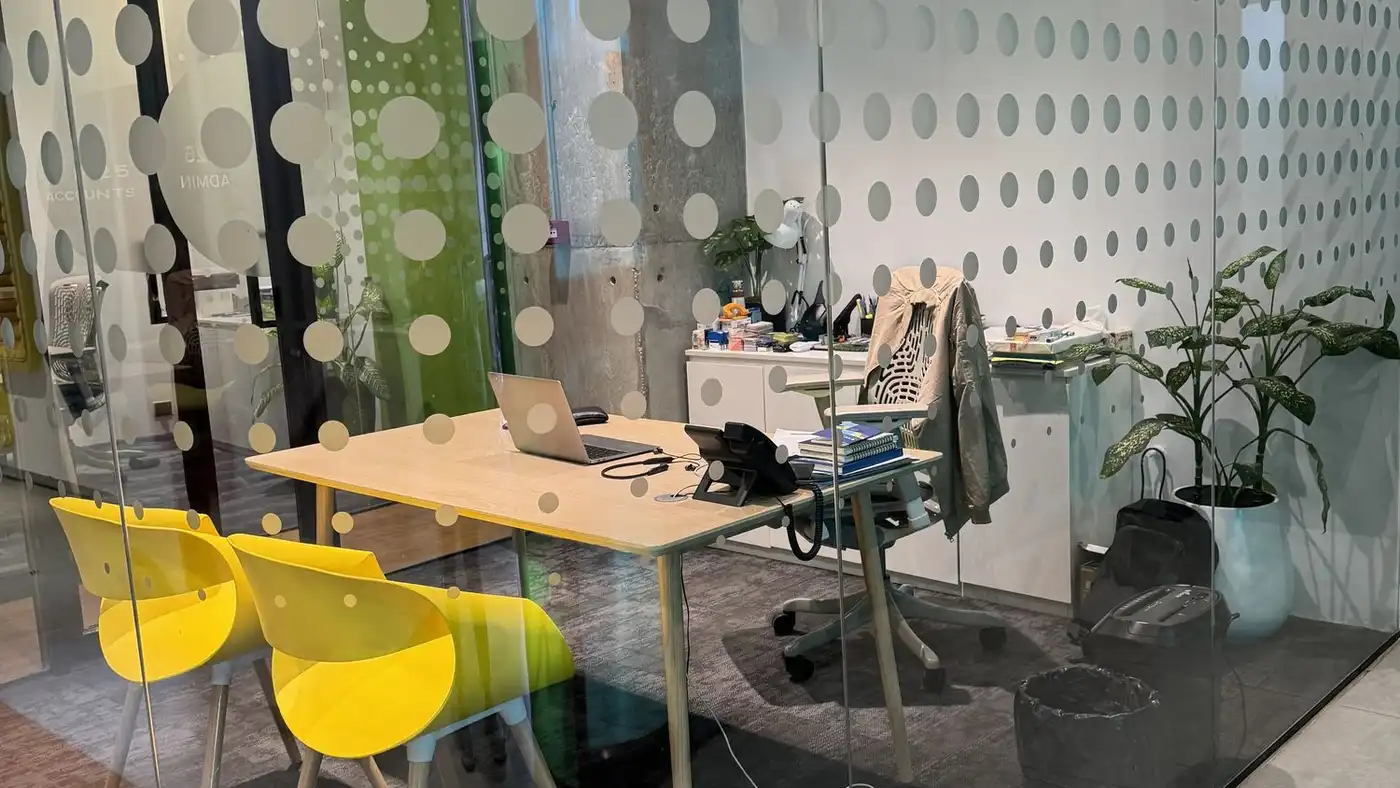 A glass-walled office with a desk, an open laptop, two yellow chairs and potted plants