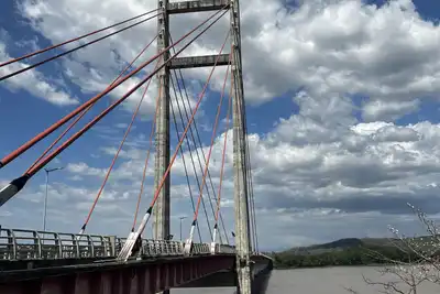 A cable-stayed bridge with red cables over a wide brown river under a blue sky with clouds