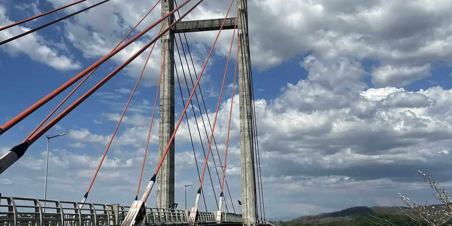 A cable-stayed bridge with red cables over a wide brown river under a blue sky with clouds