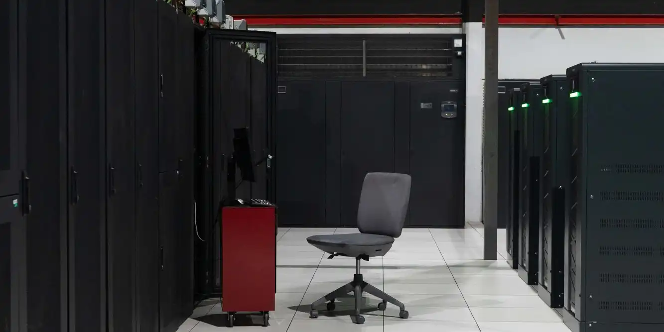 An aisle between black server cabinets in a white-floored data centre, with one office chair