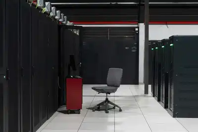 An aisle between black server cabinets in a white-floored data centre, with one office chair