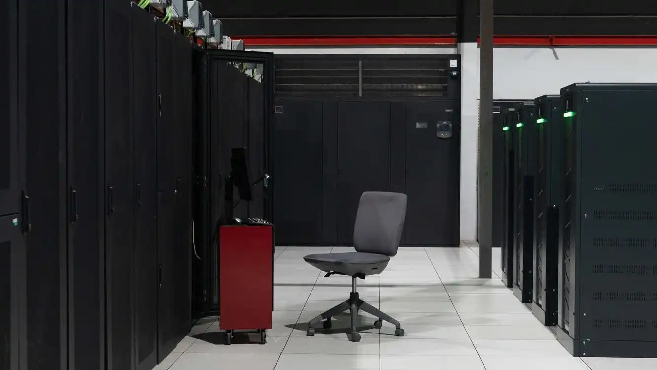An aisle between black server cabinets in a white-floored data centre, with one office chair
