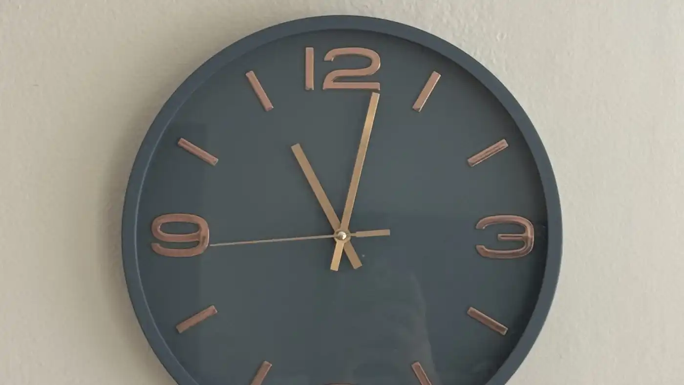 A grey wall clock with copper numerals on a plain white wall, showing about five to twelve