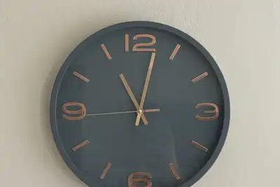 A grey wall clock with copper numerals on a plain white wall, showing about five to twelve